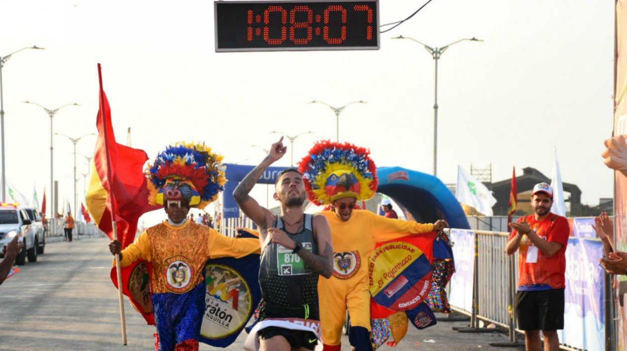 Gerard Giraldo cruza la meta escoltado por dos personajes carnavaleros. 