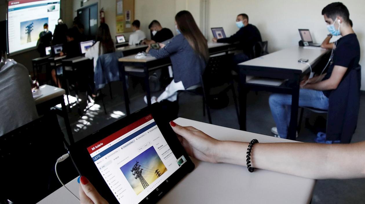 Varios alumnos siguen con ordenadores y a través de una pantalla la clase que esta impartiendo una profesora de forma presencial a otro grupo de alumnos universitarios en otro aula.
