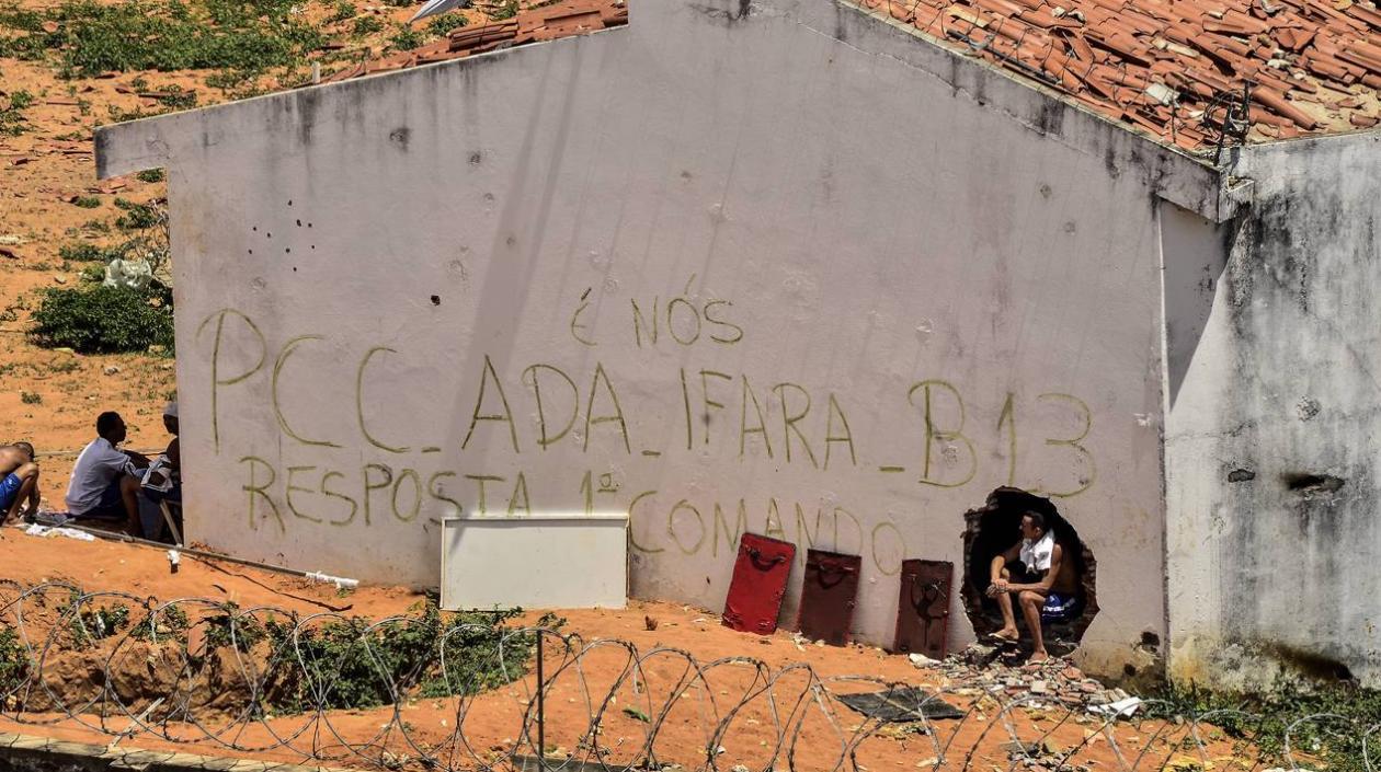 un grupo de presos junto a un muro en el que se leen las siglas del Primer Comando de la Capital (PCC)