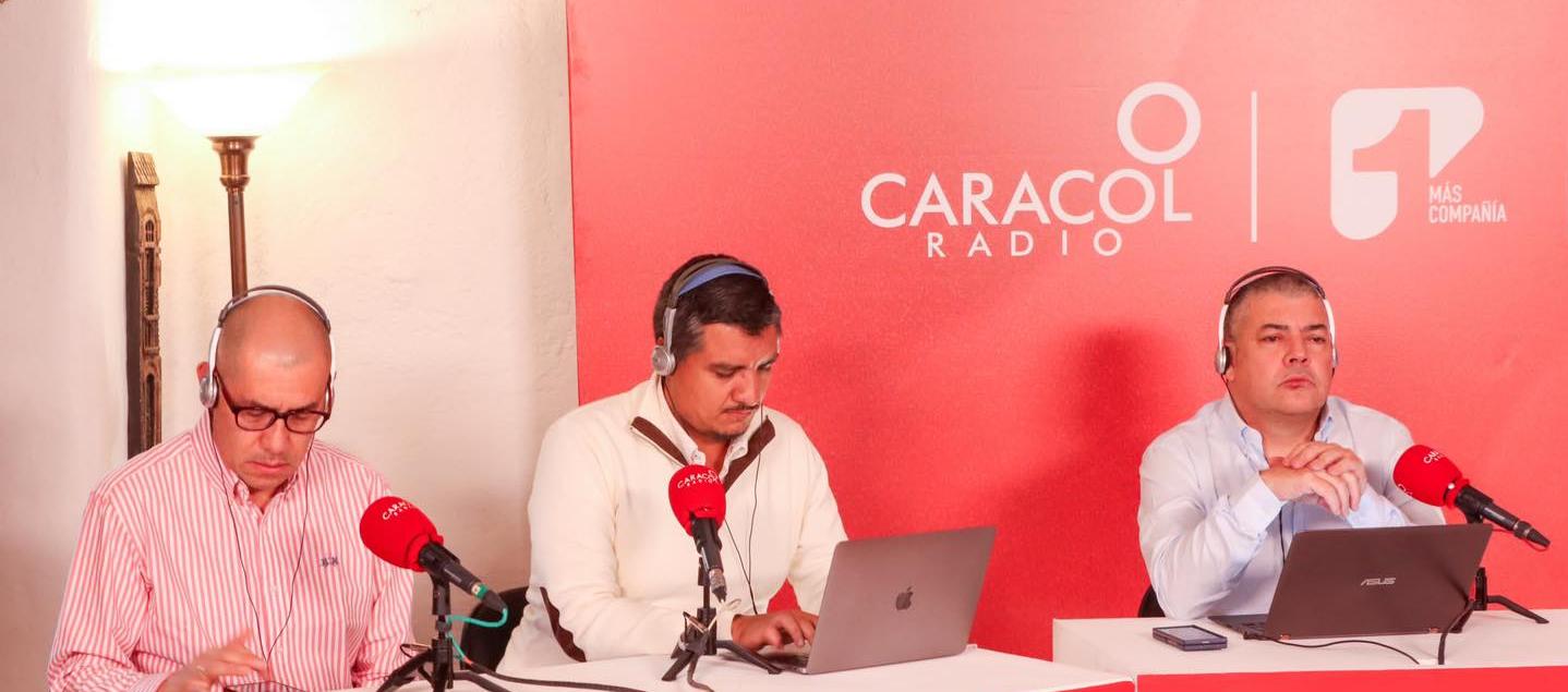 Logos de Caracol Radio y Canal 1 compartiendo espacio en una mesa de trabajo de un programa de la cadena radial.