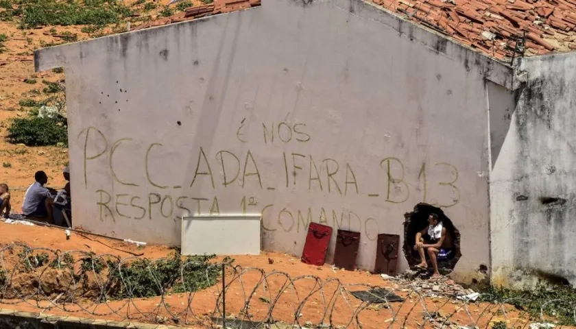 un grupo de presos junto a un muro en el que se leen las siglas del Primer Comando de la Capital (PCC)