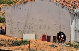 un grupo de presos junto a un muro en el que se leen las siglas del Primer Comando de la Capital (PCC)