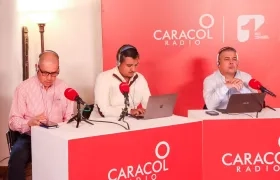Logos de Caracol Radio y Canal 1 compartiendo espacio en una mesa de trabajo de un programa de la cadena radial.