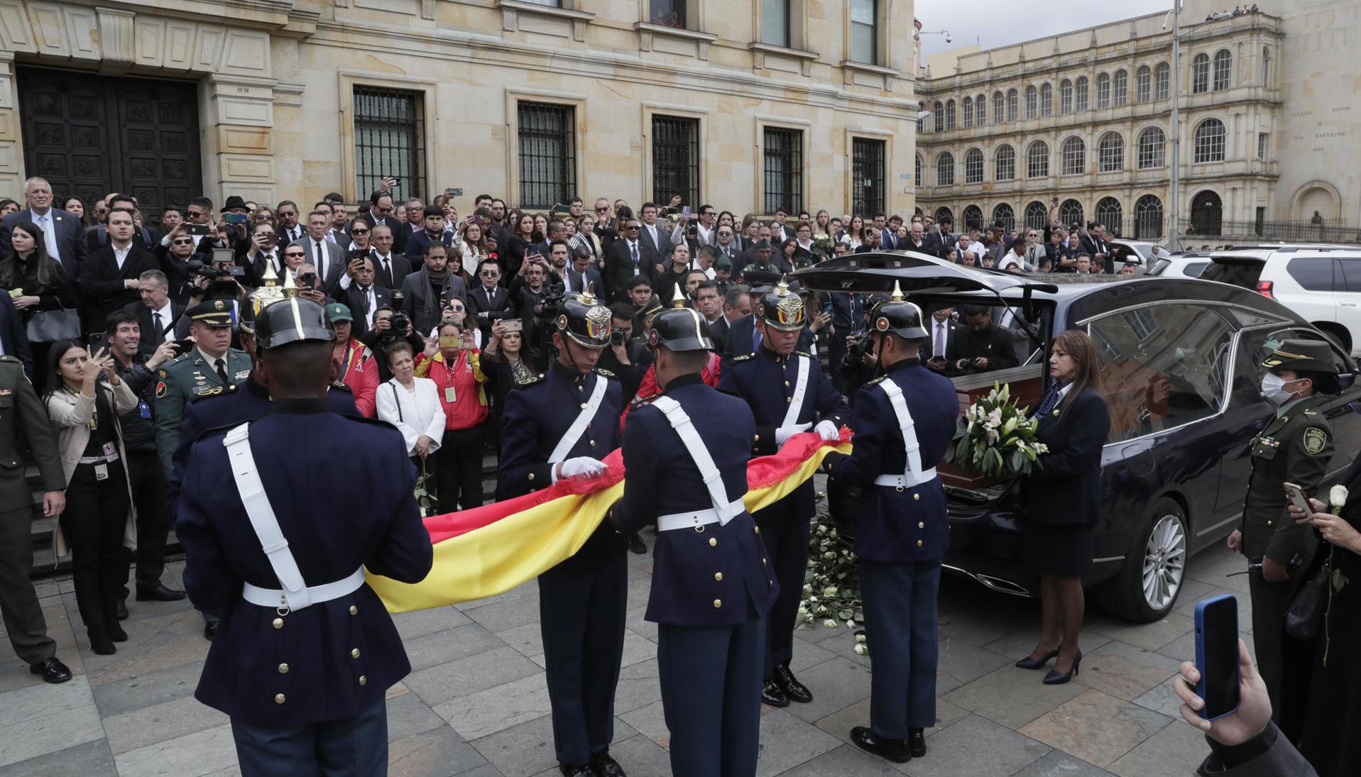 Integrantes del Batallón Guardia Presidencial doblan una bandera de Colombia junto a coche fúnebre con el féretro de Miguel Uribe.