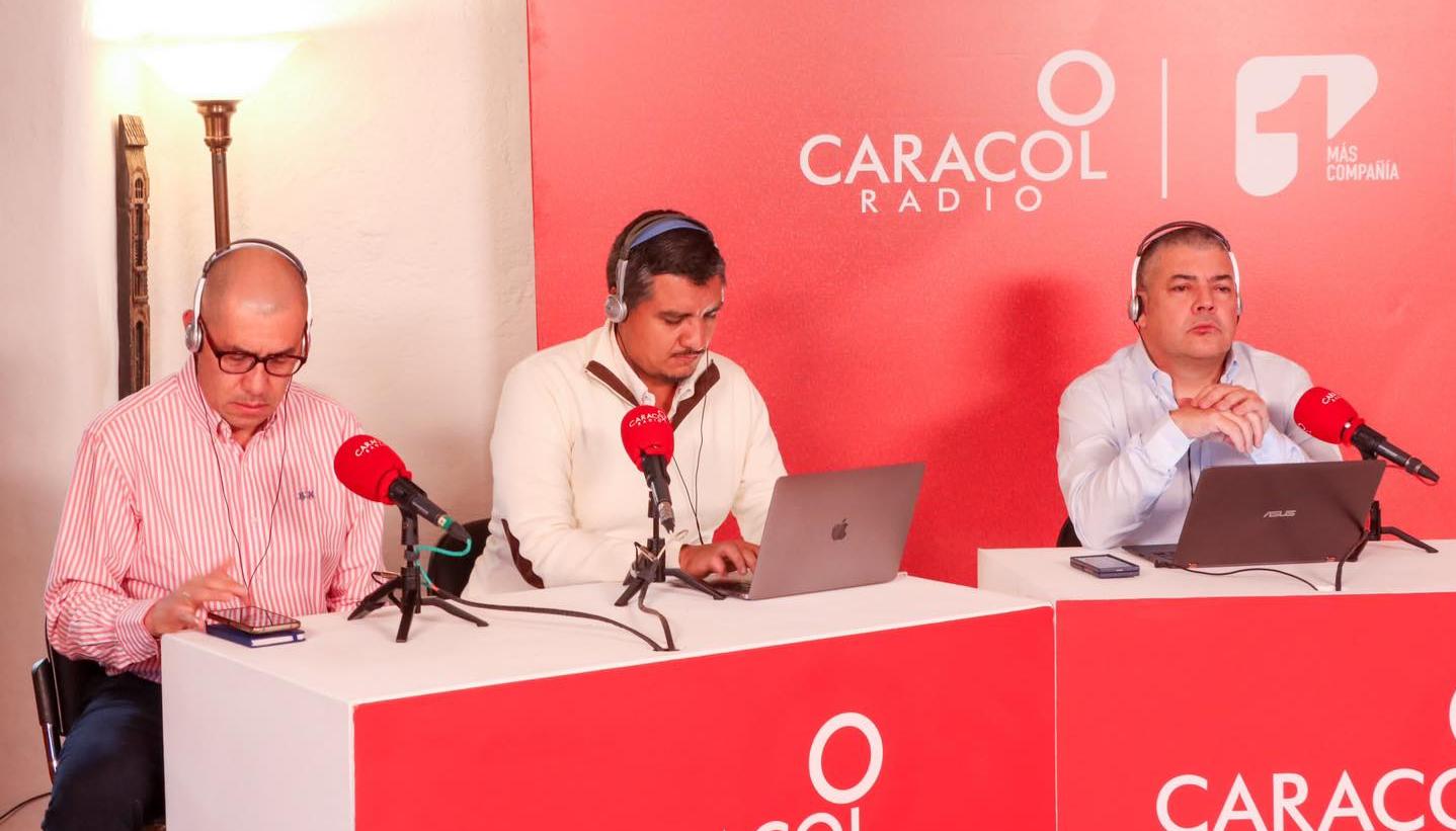 Logos de Caracol Radio y Canal 1 compartiendo espacio en una mesa de trabajo de un programa de la cadena radial.