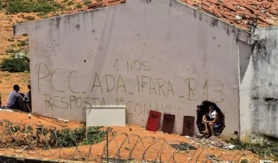 un grupo de presos junto a un muro en el que se leen las siglas del Primer Comando de la Capital (PCC)